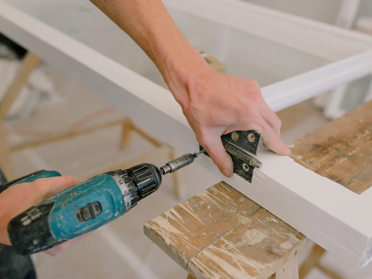 Hands driving a screw into a metal hinge on a white wooden frame with a cordless drill.