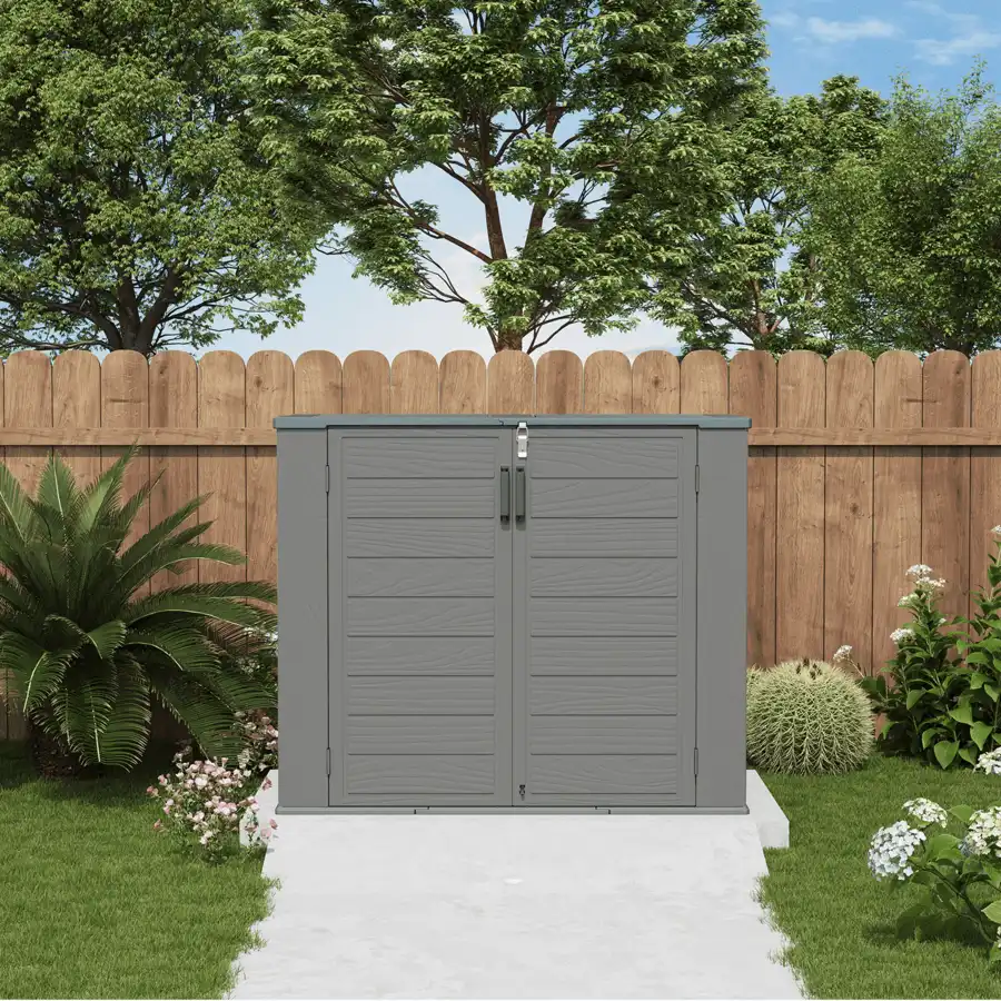 Gray resin storage cabinet with two doors standing against a wooden backyard fence.